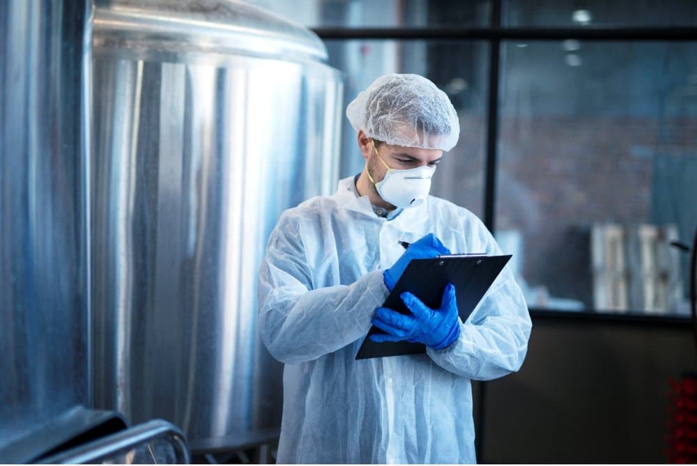 A technician in cleanroom clothing taking notes beside a stainless steel production vessel
